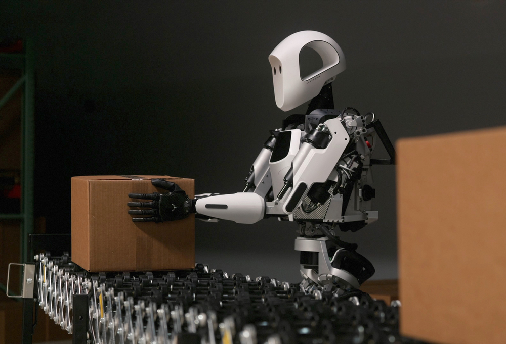 A human-like robot places a cardboard box on a conveyor belt.